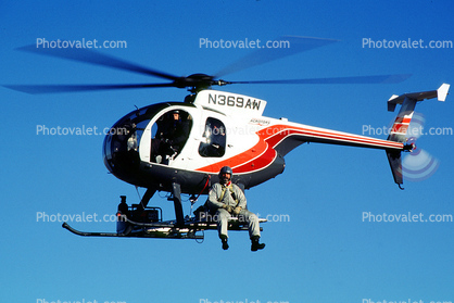 N369AW, Hughes 369D, ACROTORS, Man sitting on a skid in flight