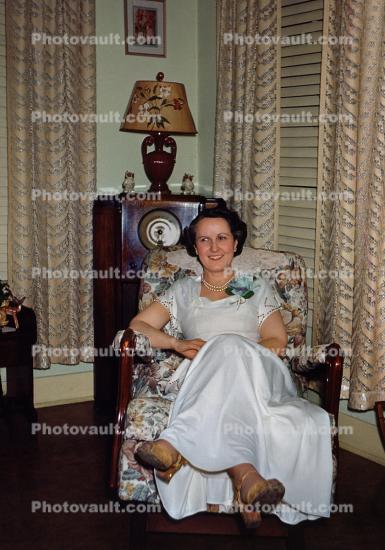Woman sits, formal dress, lamp, lampshade, radio, drapes, curtains, smiles, corsage, 1940s