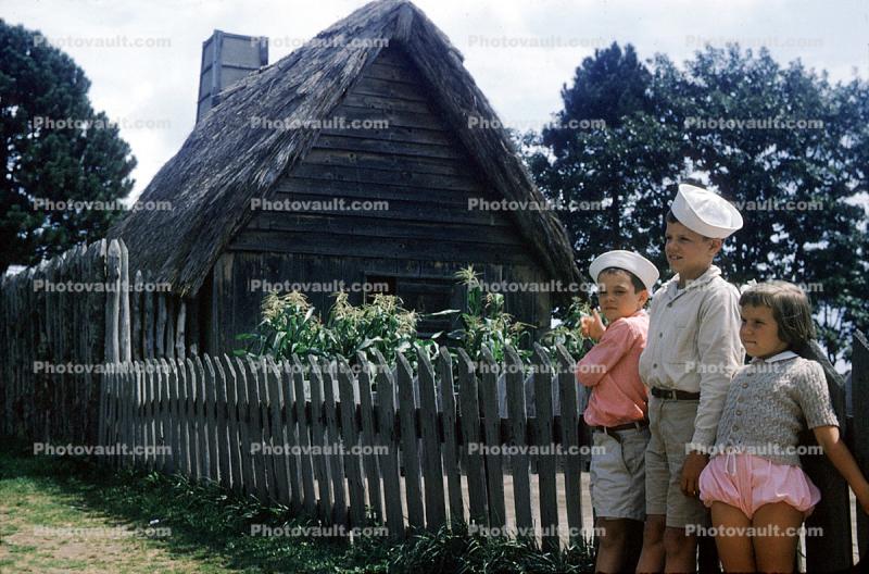 Boys, girl, fence, grass roof, house, building, 1950s