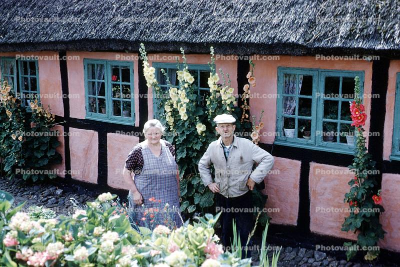 Woman and Man in front of their house, thatched roof, flowers, 1950s, Sod