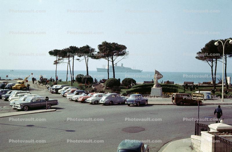 Cars in a parking lot, trees, ship, statue, 1950s