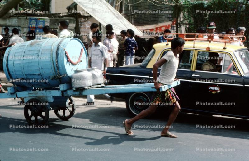 Man pulling on a cart, water, barrel, taxi cab, car, on the Streets of Mumbai